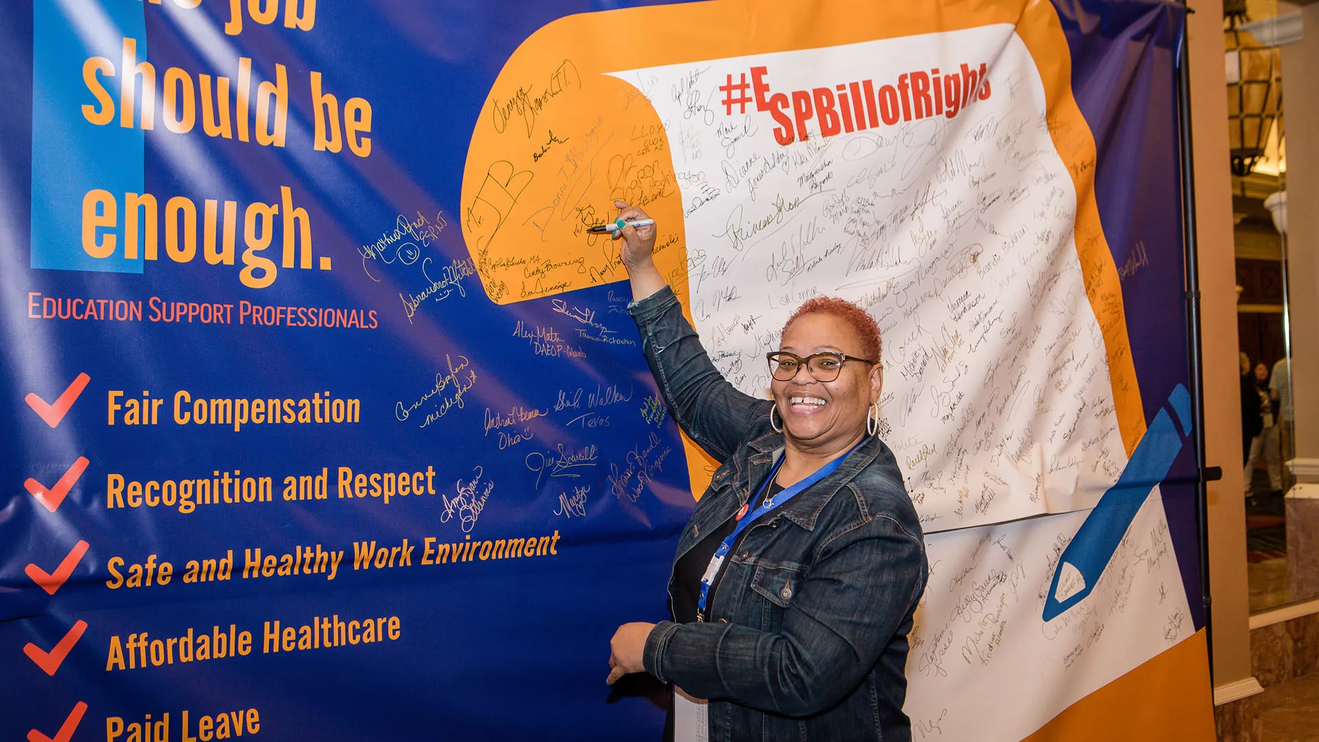 a photo of an education support professional holding a pen up to sign a large yellow-outlined scroll of the ESP Bill of Rights.